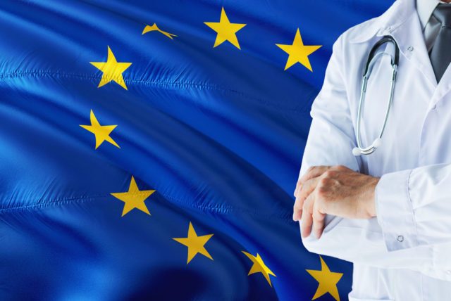 European Doctor standing with stethoscope on European Union flag background. National healthcare system concept, medical theme.European Doctor standing with stethoscope on European Union flag background. National healthcare system concept, medical theme.E